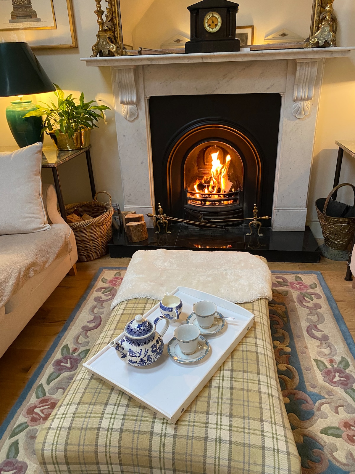 A cozy living area features a classic fireplace with an inviting flame. A large, light-colored sofa sits nearby, complemented by a decorative area rug. A tray holds a tea set, including a teapot and cups, atop a checkered ottoman.
