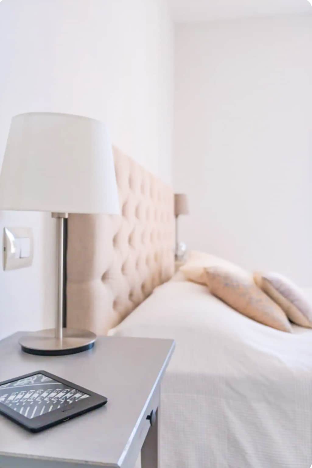A well-lit sleeping area features a soft headboard and neatly arranged pillows on a spacious bed. A bedside table holds a tablet and a sleek lamp, contributing to a minimalist design. Light-colored walls enhance the room's bright atmosphere.