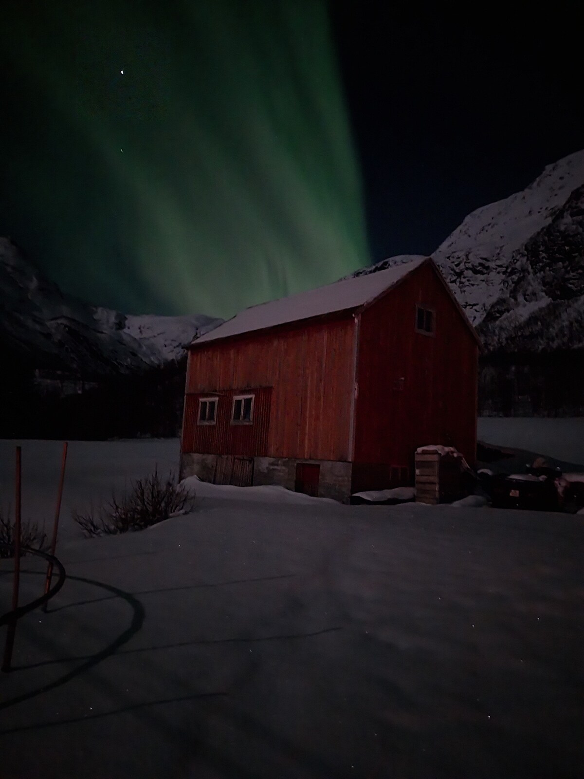 A charming red wooden house is set against a backdrop of snow-covered ground and towering mountains. The house reflects the stunning display of the Northern Lights above, casting a soft green hue. The scene is serene, emphasizing the peacefulness of the surrounding wilderness.
