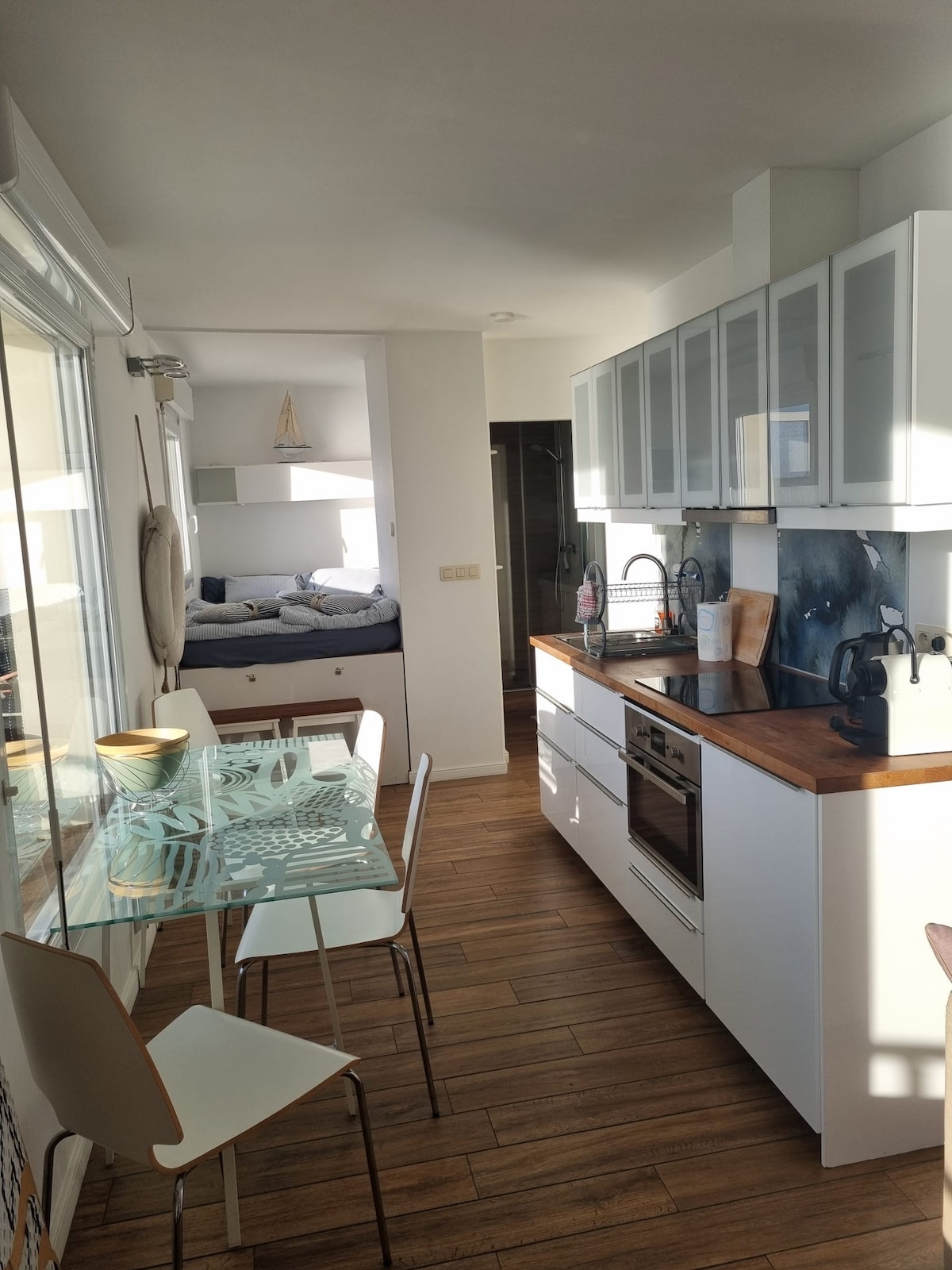 A small, fully equipped kitchen features modern white cabinetry and a wooden countertop. A glass dining table with four chairs is present. Light floods the space through large windows, illuminating the open layout connecting to the sleeping area, which is partially visible in the background.