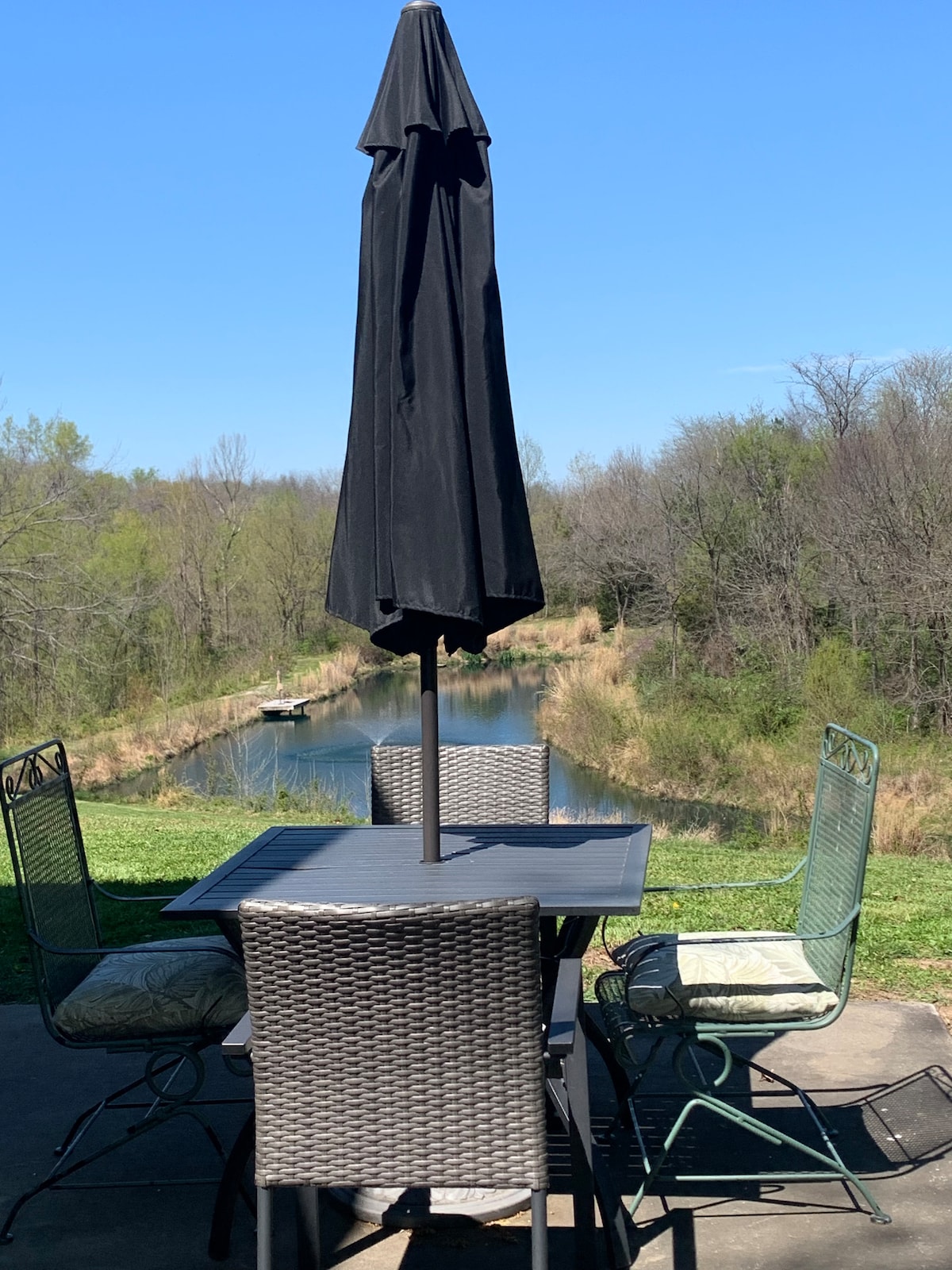 A patio table with four chairs and a large black umbrella provides a serene outdoor dining area. Lush greenery frames the view of a calm pond in the background, contributing to a tranquil atmosphere.