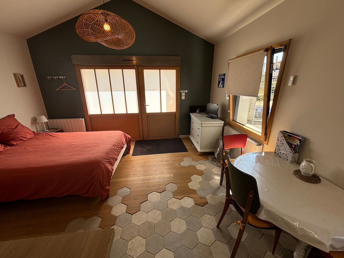A bright and welcoming studio space is presented, featuring a spacious bed with earthy tones. A small dining area with a round table and chairs is visible. A desk and computer provide a workspace, while large glass doors allow natural light to fill the room.