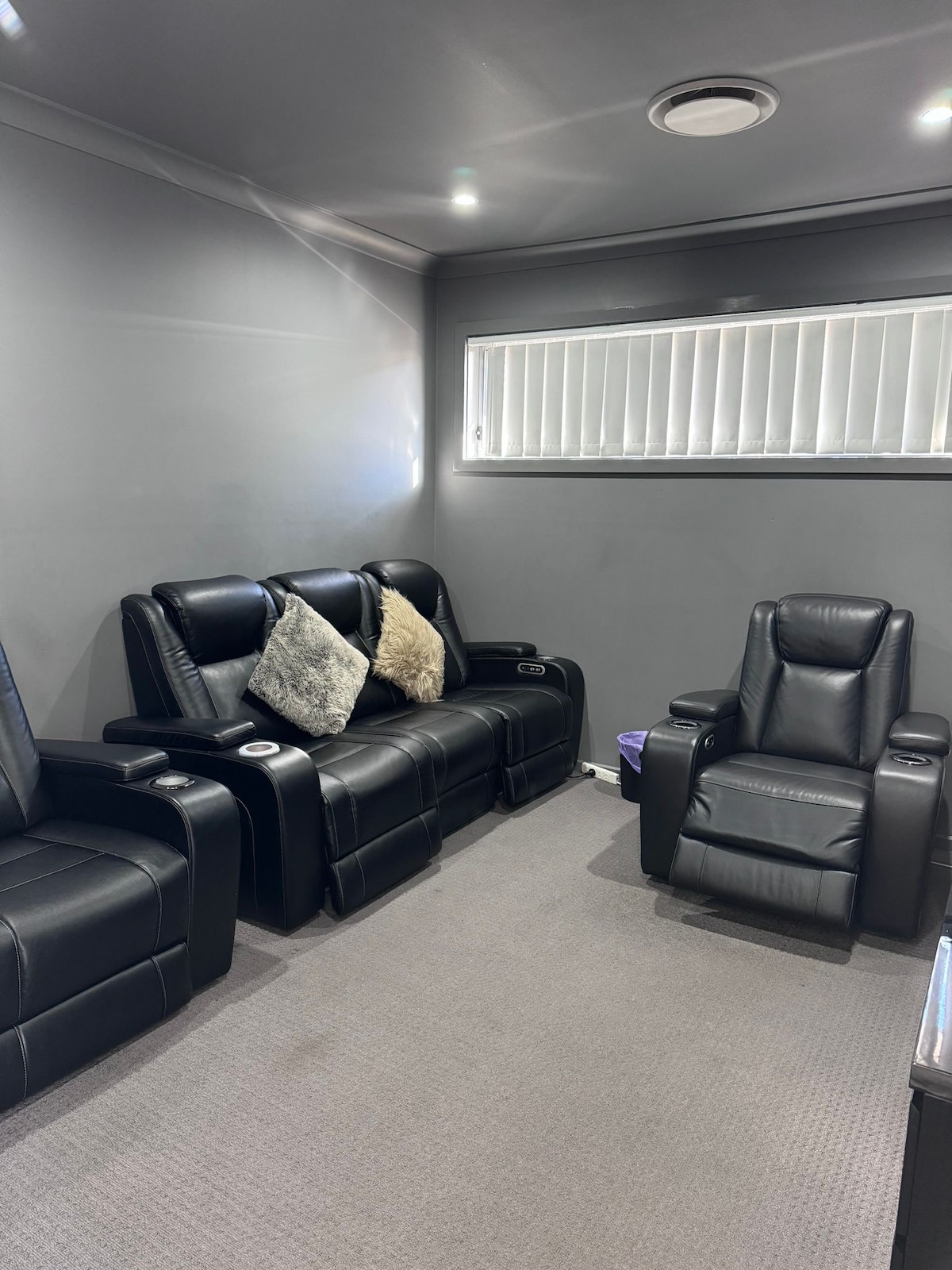 A designated movie room contains five recliner seats upholstered in black leather, arranged to provide comfort during film viewings. Natural light is filtered through a set of horizontal blinds on the window. A plush carpet covers the floor, enhancing the cozy environment.