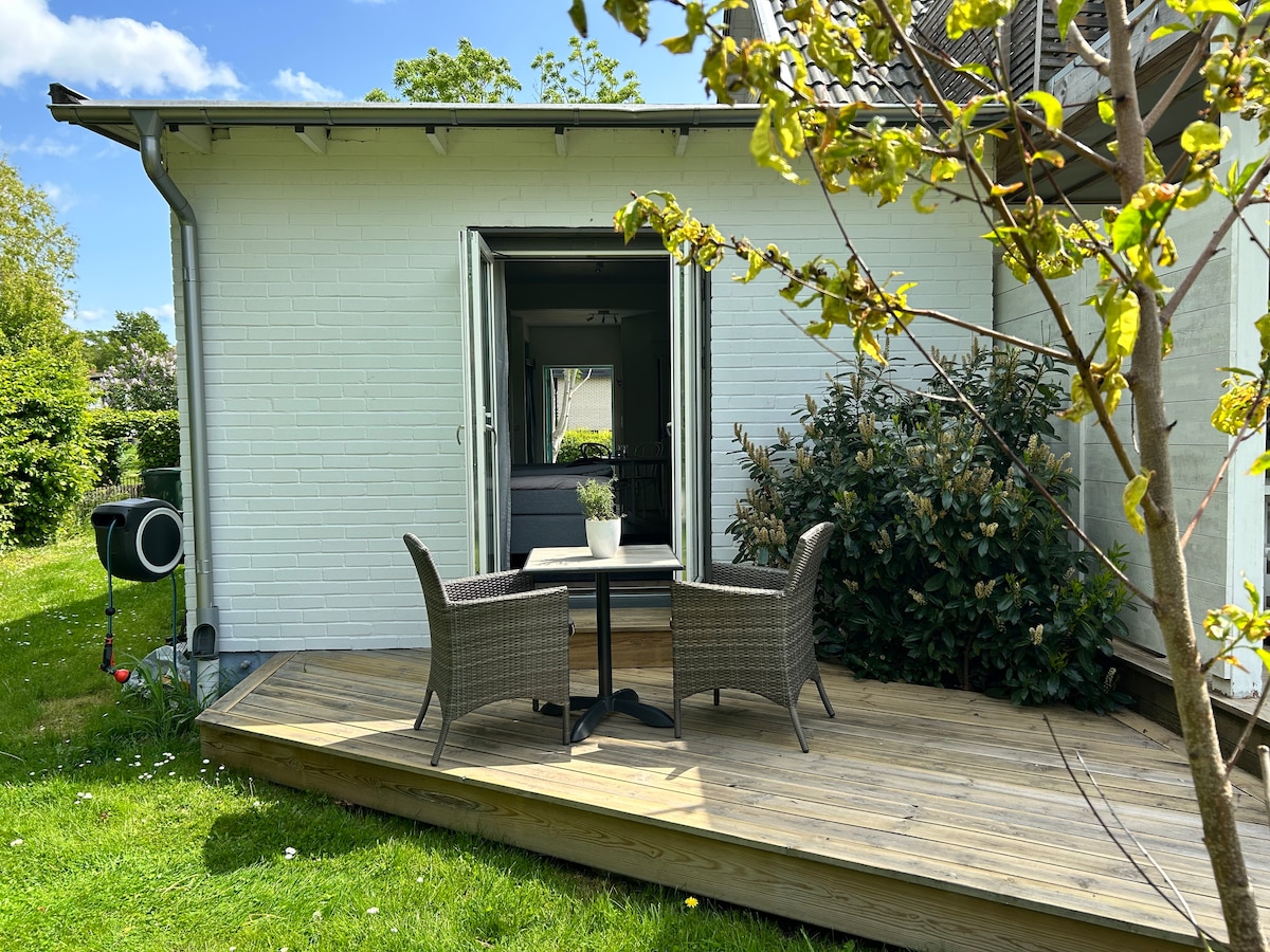 A wooden deck is shown, featuring two woven chairs and a small round table. Green grass surrounds the area, and a shrub with lush green foliage is nearby. A doorway is visible, leading into the interior space.