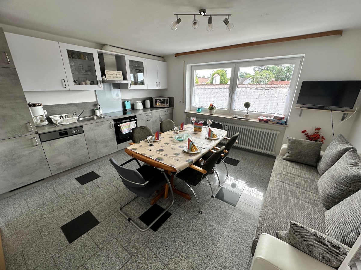 An open kitchen area features a dining table set for six with decorative tableware. Kitchen appliances, including a toaster and microwave, are neatly arranged. A cozy seating area with a sofa is positioned alongside a window that allows ample natural light to fill the space.