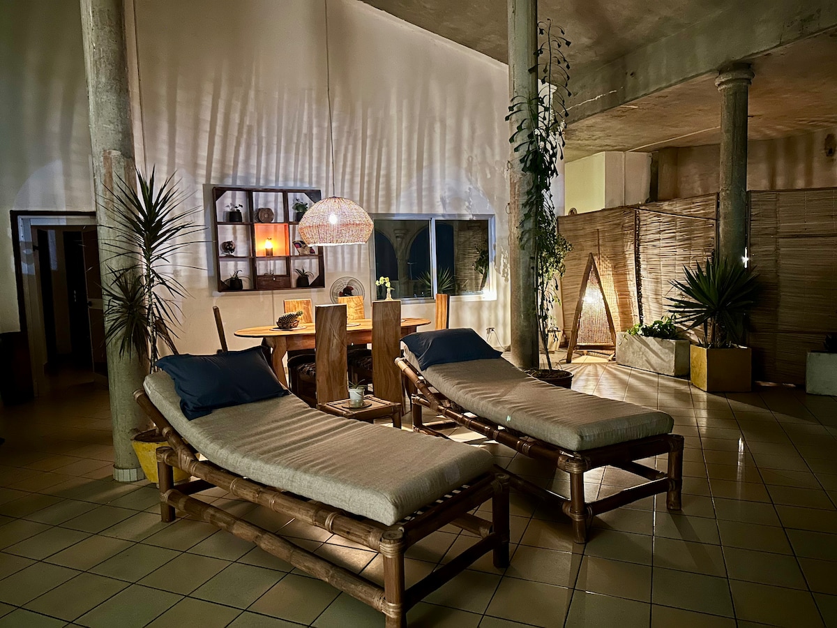 Two comfortable lounge chairs are positioned next to a table in a spacious living area. Soft lighting creates a relaxed ambiance, while plants add a touch of nature. A dining area is visible in the background, complemented by unique decorative elements along the walls.