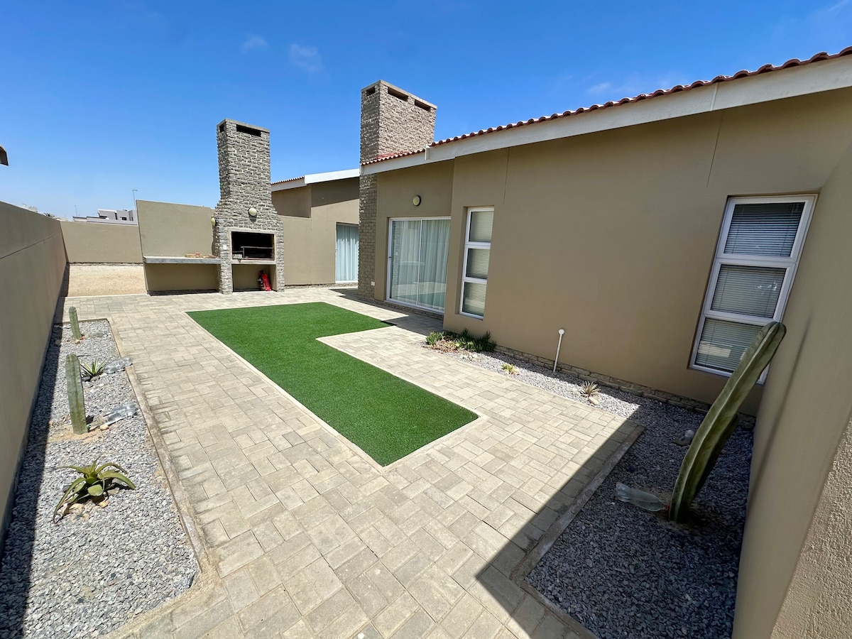 An outdoor area features a paved terrace with artificial grass and an arrangement of low-maintenance plants. Two stone chimneys stand beside a built-in barbecue area, while light-colored walls create an open and airy ambiance. Clear skies suggest a bright day.