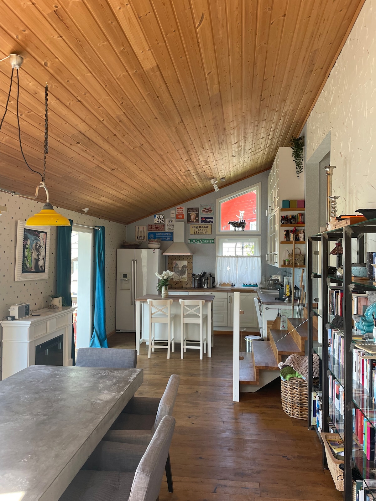 A well-lit interior features a modern kitchen with light cabinetry and a dining area with a gray table and four chairs. A cozy seating area is highlighted by a fireplace and decorative shelving. Pine wood ceilings add warmth to the open space.