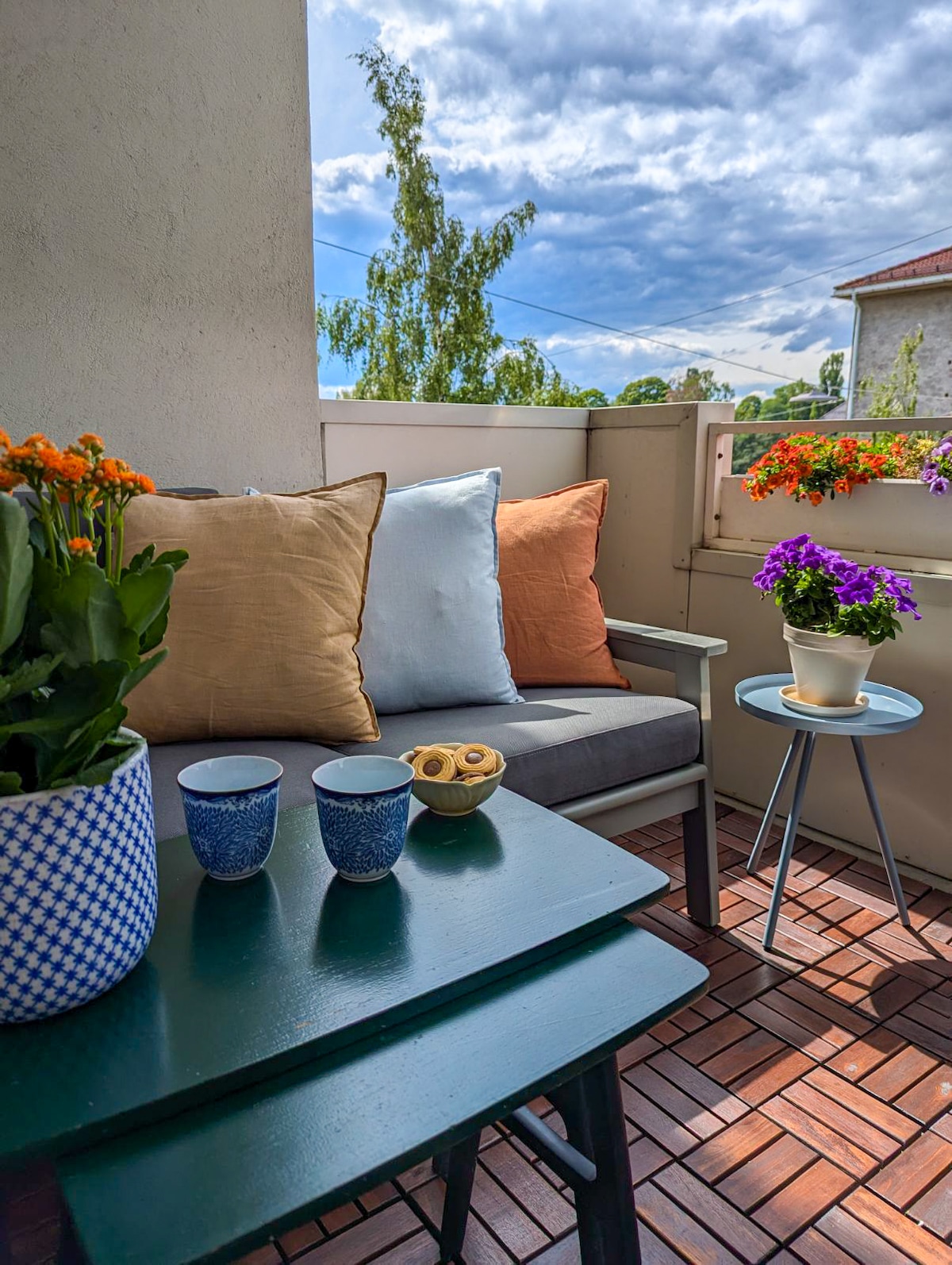 A cozy balcony setting is featured with two cushioned chairs and a small table. A potted plant and colorful flowers add charm, while a bowl of snacks rests on the table. The surrounding area is illuminated by natural light, highlighting the inviting space.