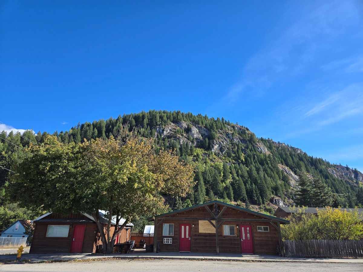 The exterior of a rustic wooden building is framed by lush greenery and towering hills in the background. A large tree offers shade in the front, while two separate entrances are visible beneath the pitched roof, enhancing the inviting appearance of the property.