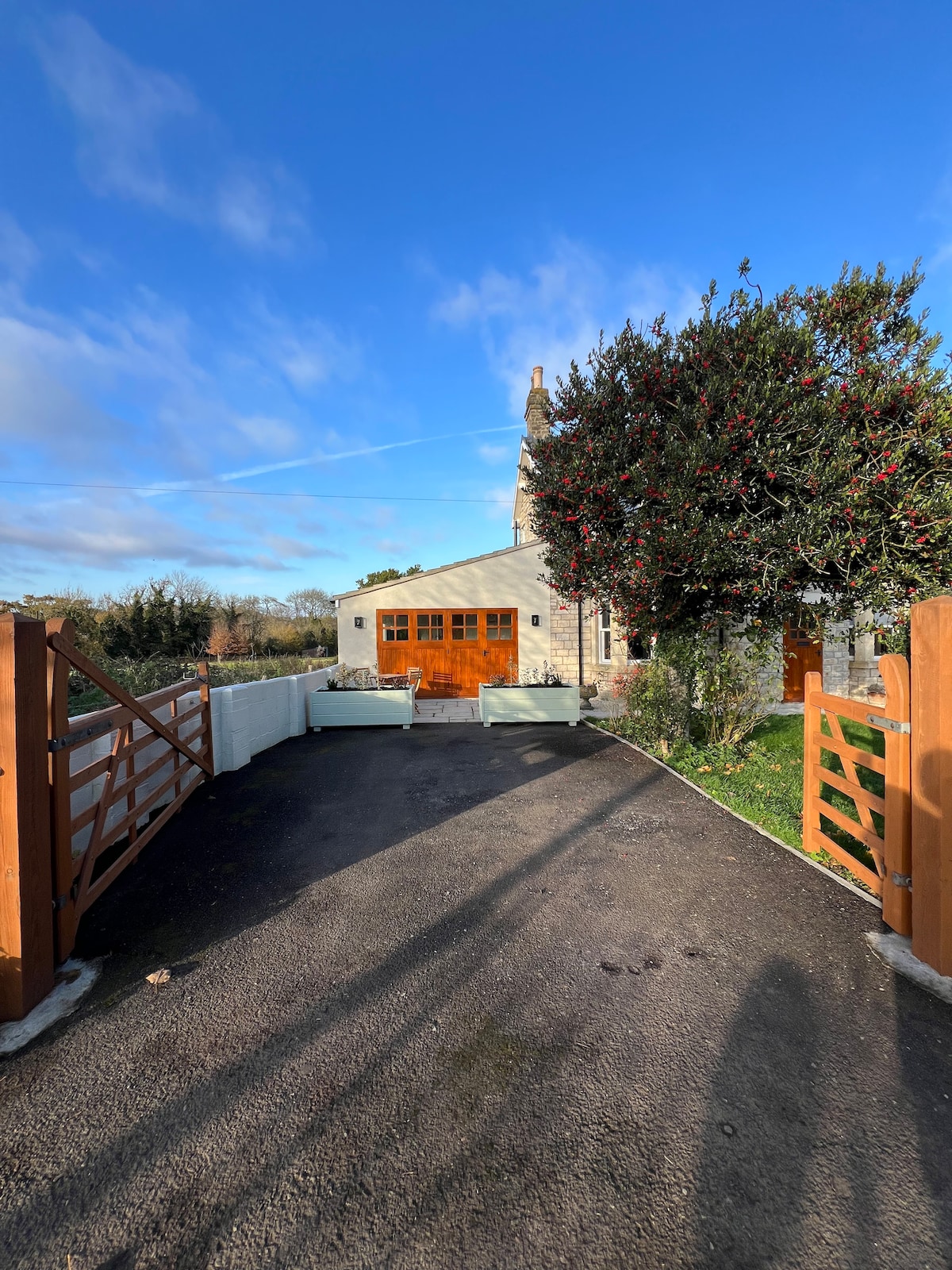 The entrance to The Old Workshop is presented, featuring a private driveway leading to a welcoming facade. Double wooden doors are visible at the end of the path, surrounded by a well-maintained landscape of greenery and trees under a clear blue sky.