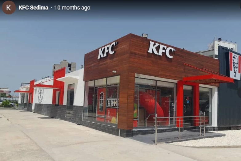 The exterior of a fast-food restaurant features a modern design with a combination of wood and white walls. Large windows provide visibility into the dining area, while a red canopy extends over the entrance. A wheelchair-accessible ramp leads to the entrance.