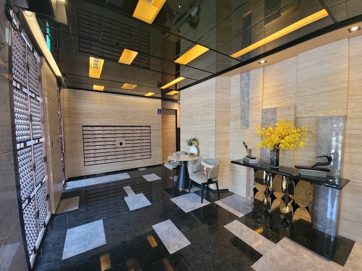 A modern lobby features polished black stone flooring and elegantly minimalistic decor. A small seating area includes two chairs and a side table. A colorful floral arrangement enhances a console table, while decorative wall details and ambient lighting create a sophisticated ambiance.