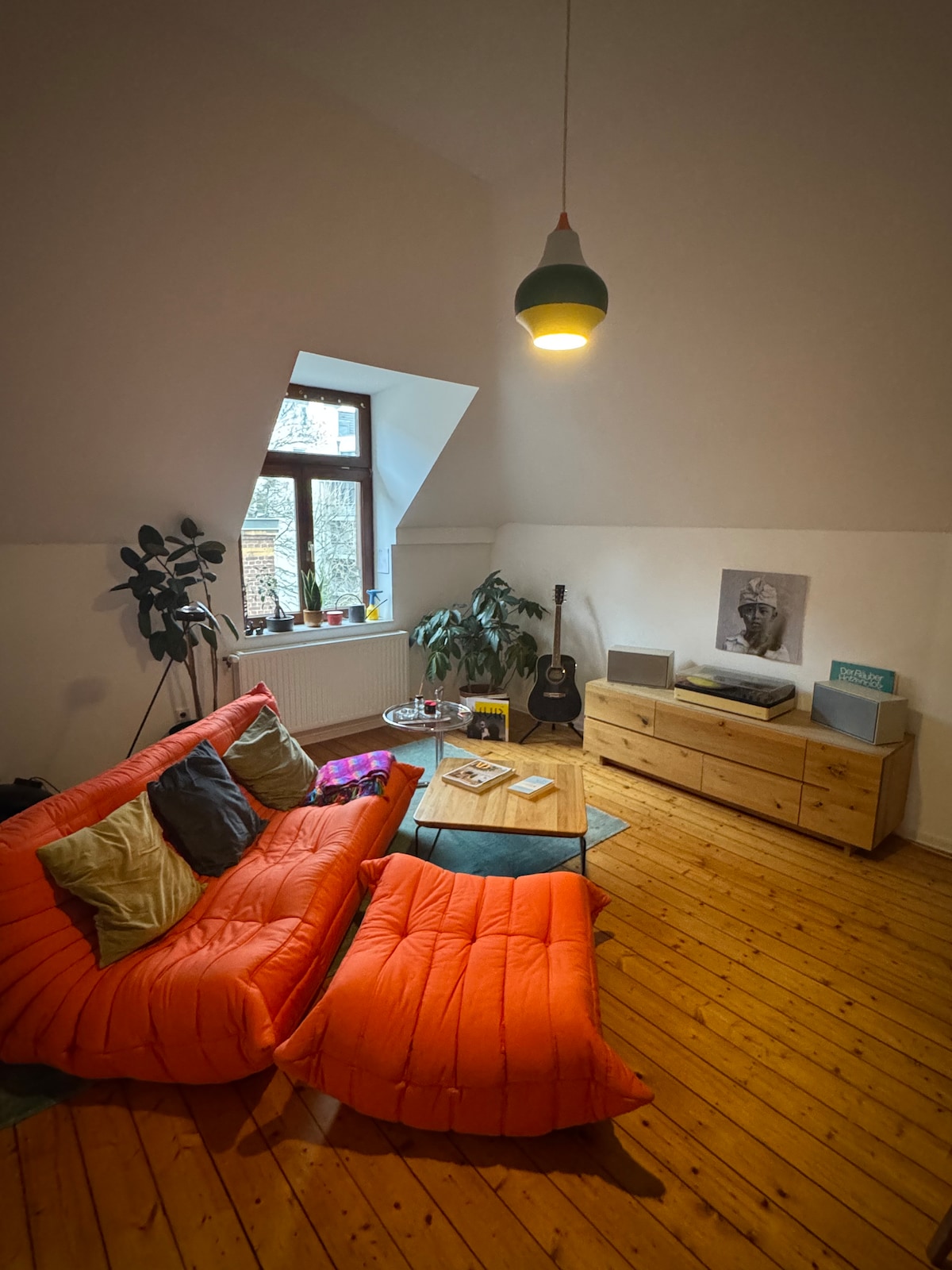 A spacious living area features a prominent orange sofa with varying cushion colors, inviting relaxation. A light wooden coffee table rests in front, complemented by a guitar in the corner. Green plants add a natural touch, while a minimalist shelving unit holds entertainment essentials.