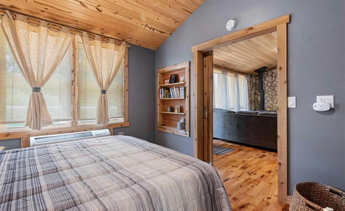 A cozy bedroom features a spacious bed covered with a plaid blanket. Large windows with textured beige curtains allow natural light to fill the room. A small bookshelf is built into the wall, and a doorway leads to a living area visible beyond.