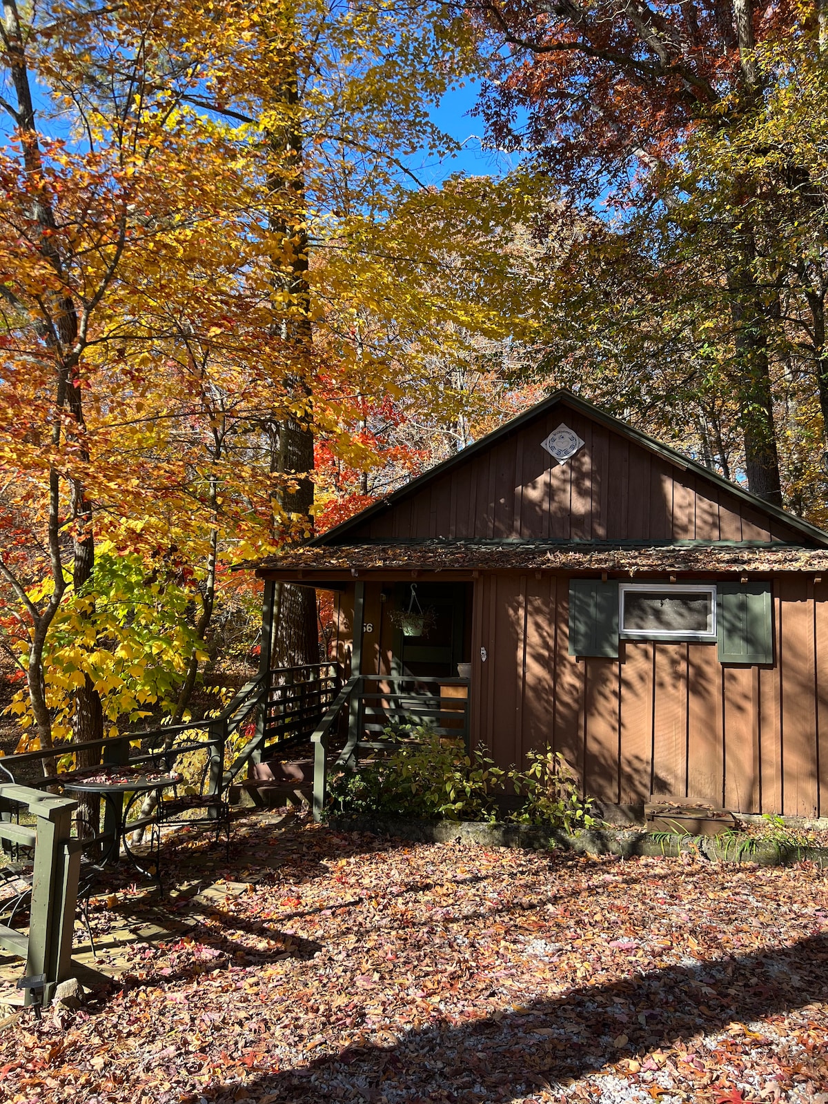The exterior of a charming cabin is surrounded by vibrant autumn foliage. A wooden deck with railing is visible, leading to the entrance. Fall leaves cover the ground, creating a colorful carpet, while the cabin features a steeply pitched roof and green shutters.