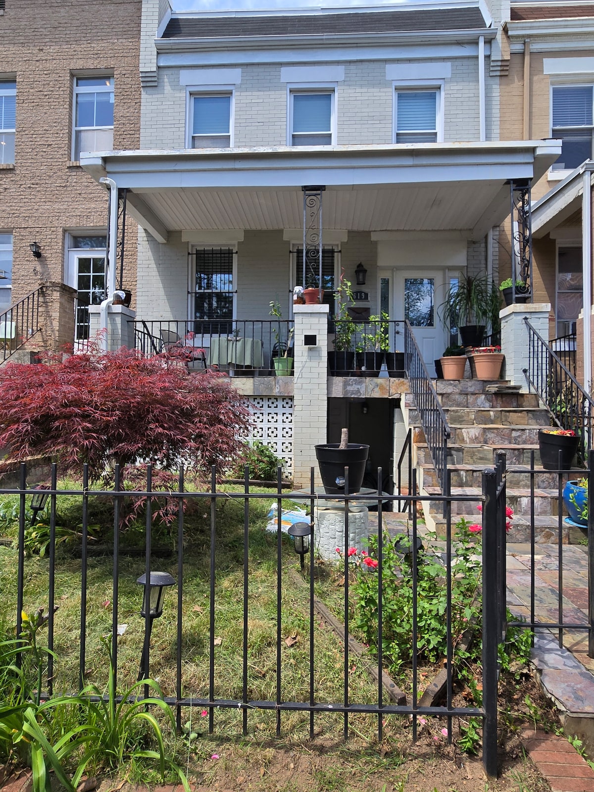 The exterior of a rowhouse is shown, featuring a front porch with plants and decorative elements. A stone pathway leads to the entrance. A garden with various flora is visible in front, surrounded by a black iron fence that encloses the space.