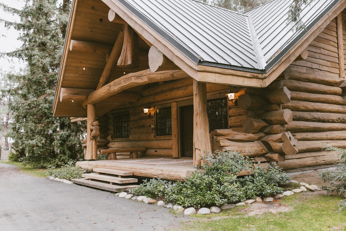 The rustic cabin exterior is constructed from large logs, featuring a sloping metal roof. A spacious porch is accentuated by wooden pillars and stone pathways, framed by lush greenery and trees, creating a serene entrance.