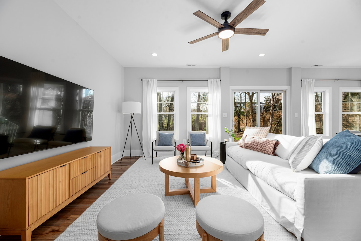 A modern living area is shown, featuring a large flat-screen TV mounted on the wall opposite a light-colored sectional sofa. A round wooden coffee table sits in the center, while two upholstered stools are positioned nearby. Natural light filters through the expansive windows adorned with sheer curtains.