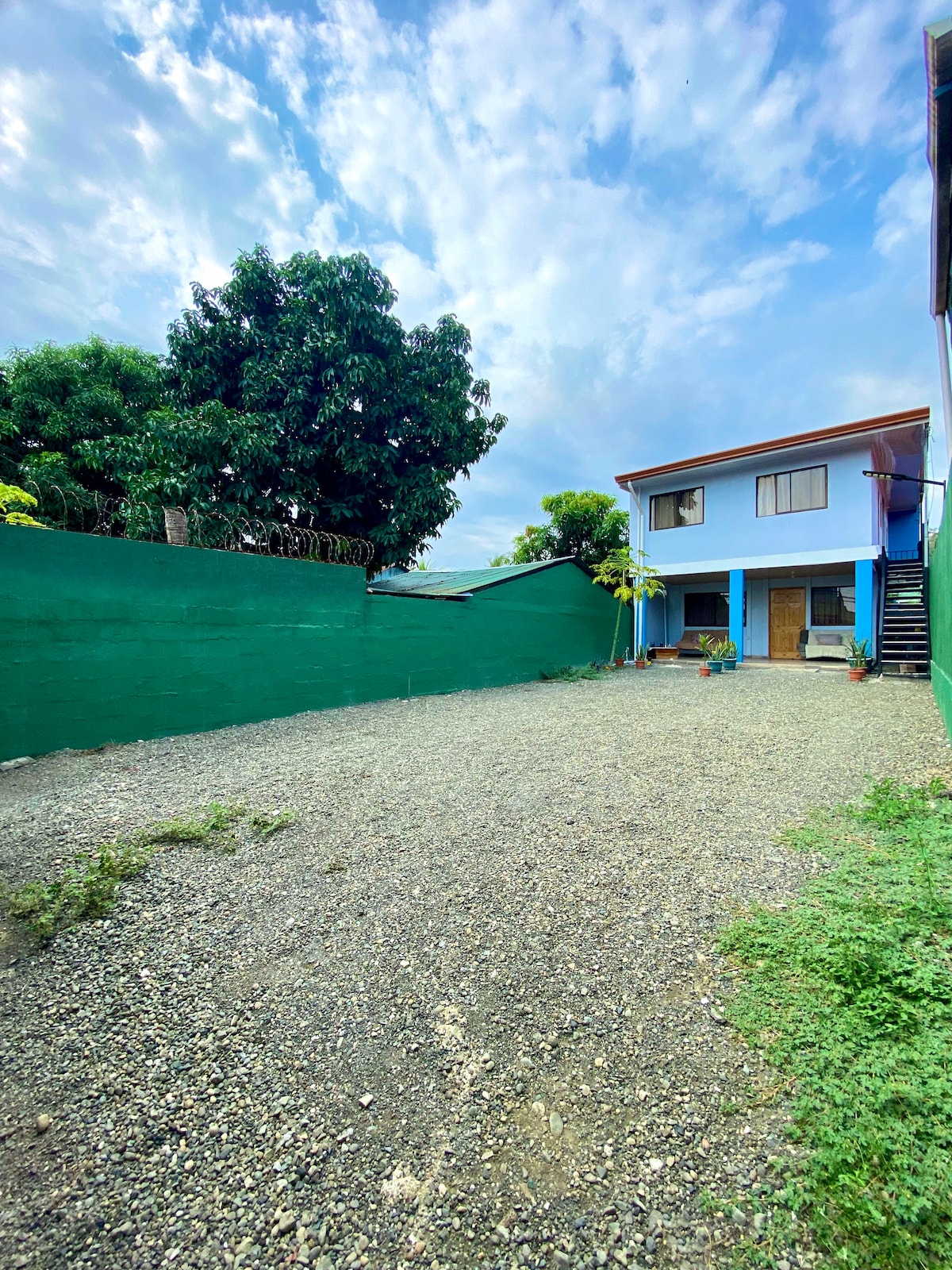 A gravel driveway leads to a two-story blue building surrounded by lush green trees. A vibrant green wall encloses the property, creating a secluded outdoor space. Several potted plants are positioned near the entrance, adding a touch of greenery to the entrance area.