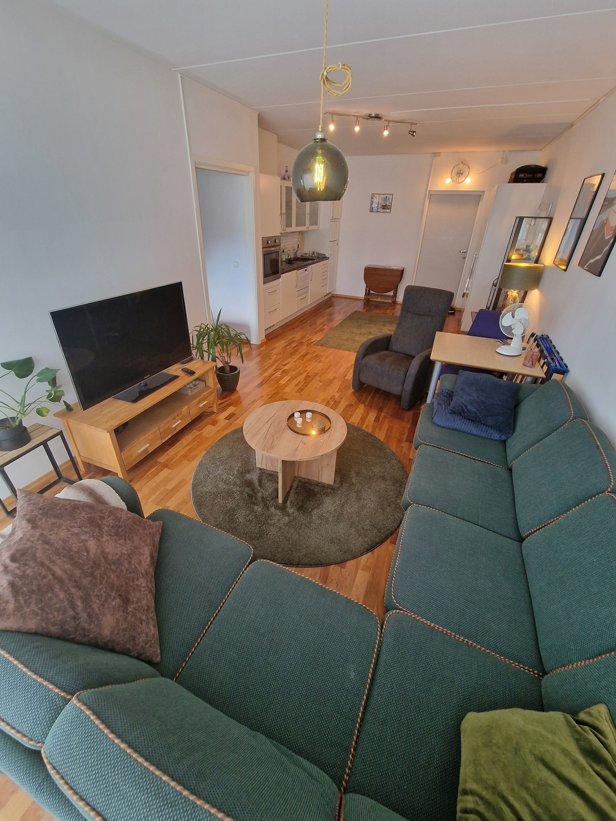 A spacious living area features a green sectional sofa and an area rug, with a round coffee table at the center. A television stands on a wooden console, while a bright kitchen area with modern appliances is visible in the background.