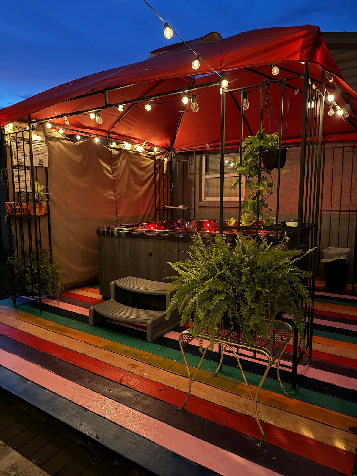 A covered hot tub area is illuminated by string lights, adding a cozy ambiance. Potted plants are positioned on shelves alongside soft drapes, and a multicolored wood deck complements the space. The enticing atmosphere invites relaxation under the evening sky.