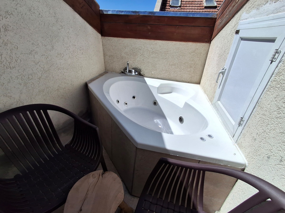 A private hot tub is positioned in a corner, featuring a smooth surface and multiple jets. Two dark, modern chairs are placed nearby on a wooden surface, providing a space for relaxation. Natural light enters through an adjacent window, highlighting the area.