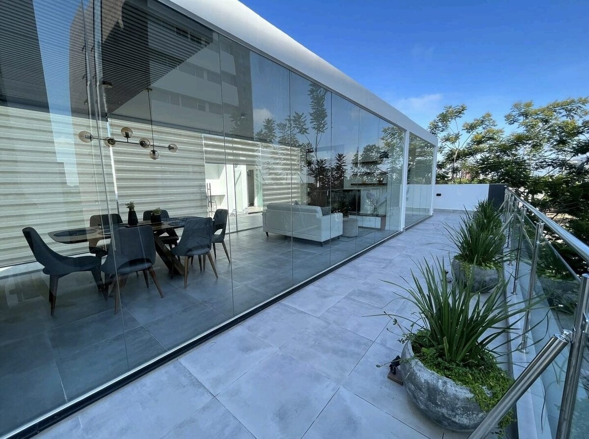 A spacious terrace is shown with modern glass walls and a view of greenery. A dining table is set with chairs, while a comfortable seating area features a light-colored couch. Potted plants add a touch of nature, contributing to a serene environment.