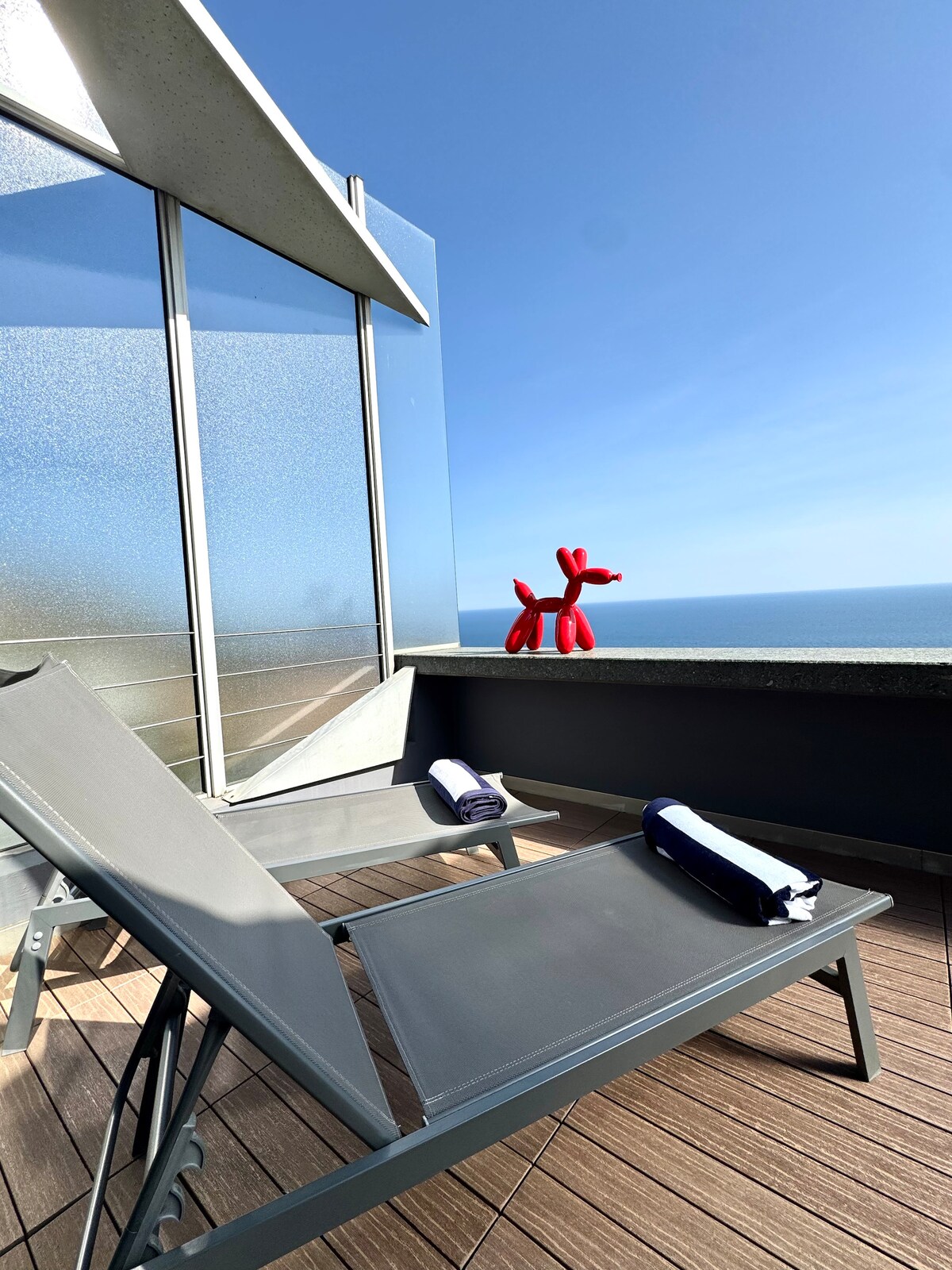 A balcony area features two gray lounge chairs, each with a towel neatly placed on the seat. A playful red balloon dog is positioned on the ledge, with a clear view of the blue sea and clear sky in the background.