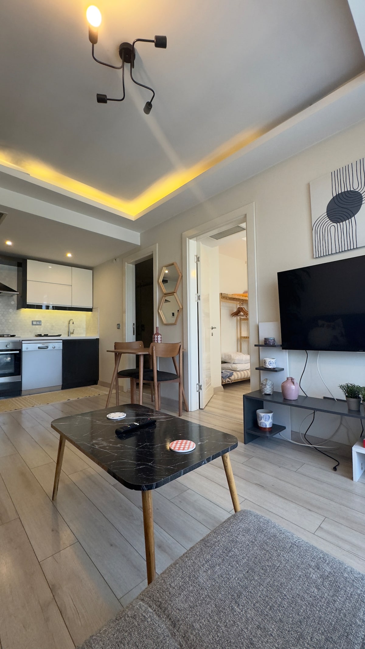 A multifunctional living space is presented, featuring a stylish coffee table and a comfortable seating area. A dining table for two is set near the kitchen. Natural light enhances the atmosphere, while a large flat-screen TV is mounted on the wall opposite the seating area.