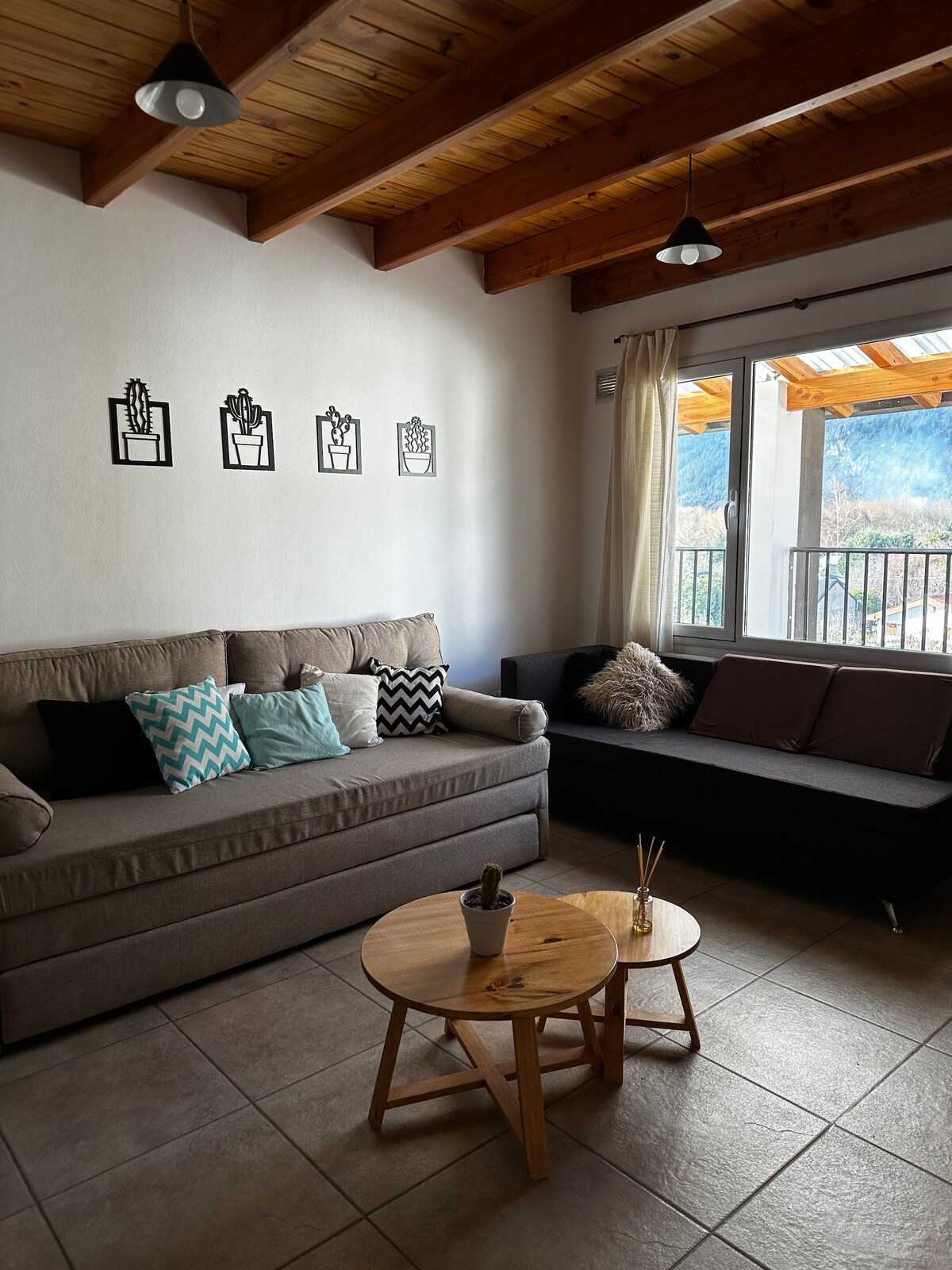 A living area is highlighted by a cozy sofa adorned with various cushions. Artistic wall decorations in simple frames hang above, and natural light enters through a large window with sheer curtains. A small wooden table sits centrally, completing the inviting space.
