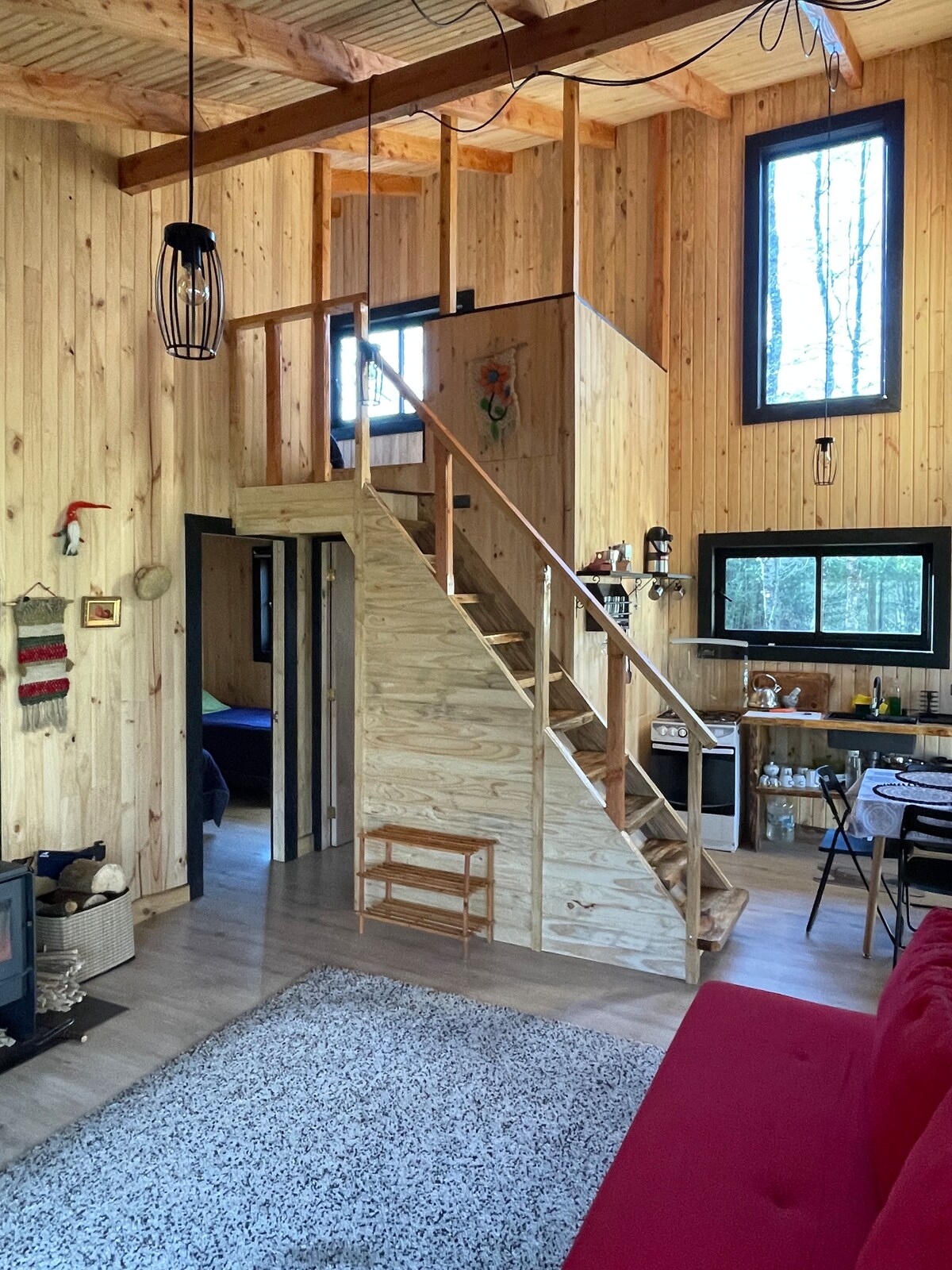 A bright interior showcases natural wood paneling and large windows allowing natural light to fill the space. A wooden staircase leads to an upper level. The living area features a red sofa and a rug, while a kitchenette is visible in the background.