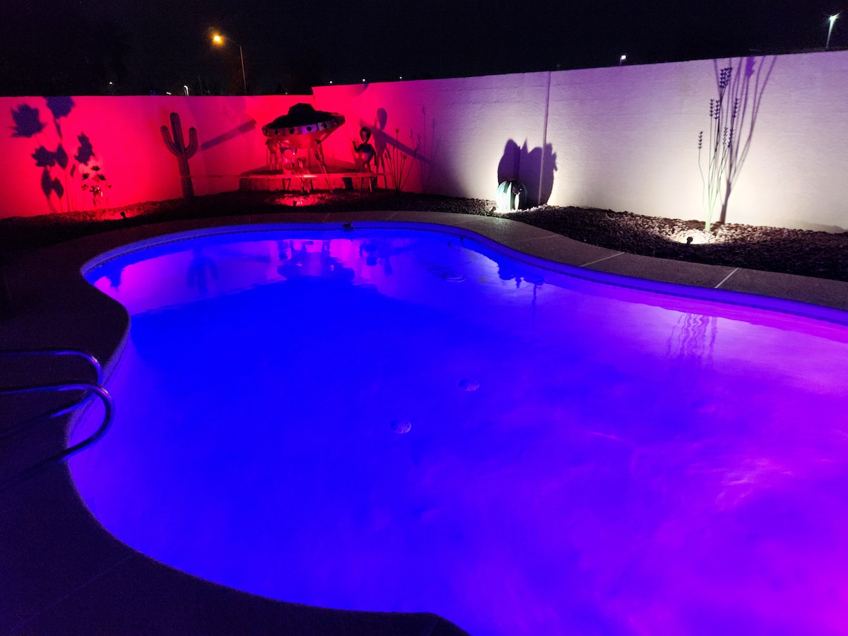 A swimming pool is illuminated with vibrant purple lighting, creating a serene atmosphere at night. The pool's shape is curvy, complementing a landscaped area that features a decorated wall with a mural, surrounded by desert-themed plants. Ambient lighting enhances the overall visual appeal.