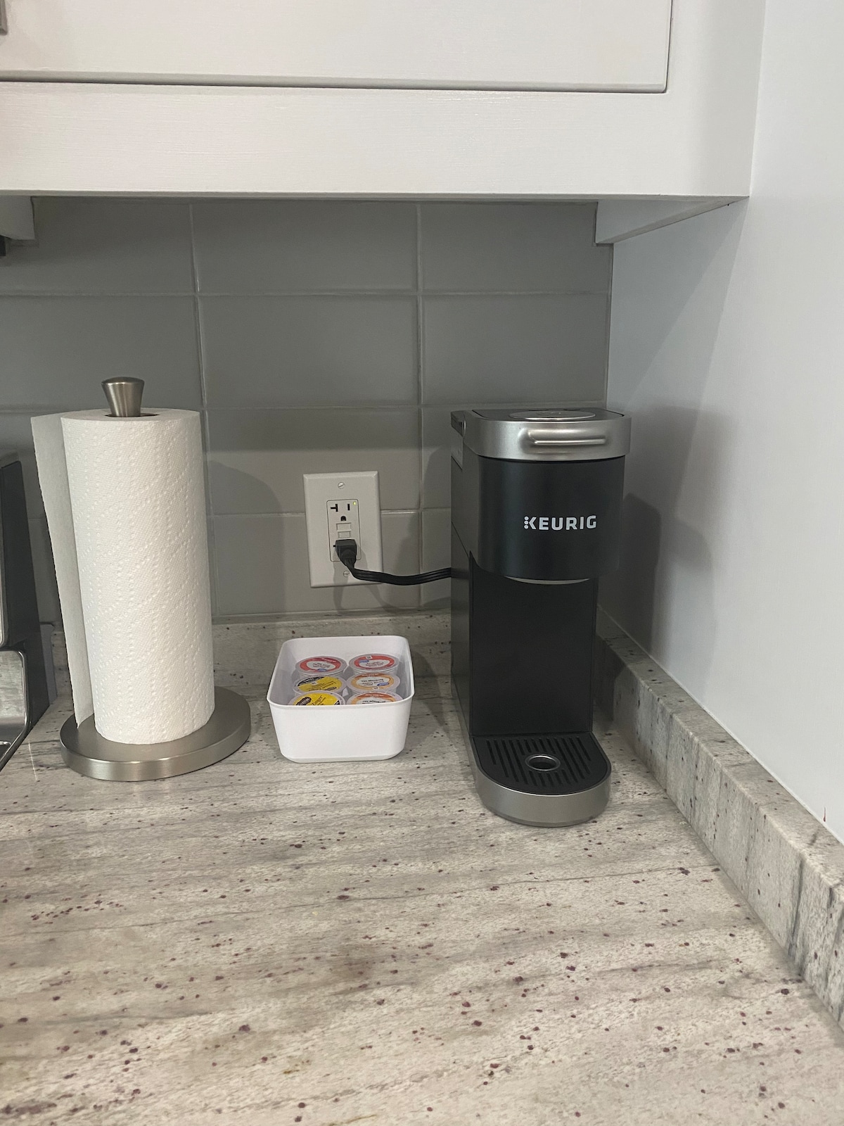 A Keurig coffee maker is positioned on a countertop, alongside a container holding coffee pods. A paper towel holder is visible, and the countertop is finished with a subtle texture. The backsplash features light grey tiles, contributing to the kitchen's modern aesthetic.