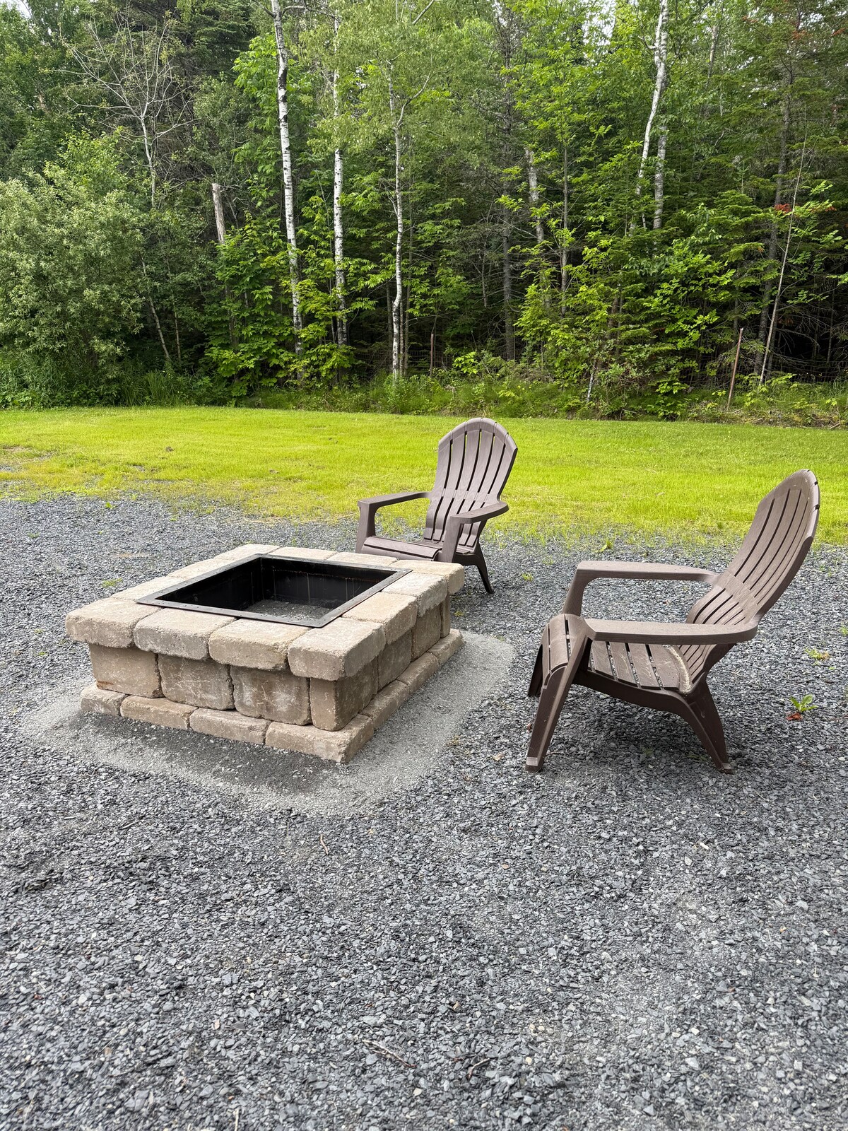 An outdoor fire pit is surrounded by a gravel area, featuring a square stone structure and two Adirondack chairs positioned comfortably nearby. Lush green trees provide a natural backdrop, enhancing the outdoor space for gatherings or quiet evenings.