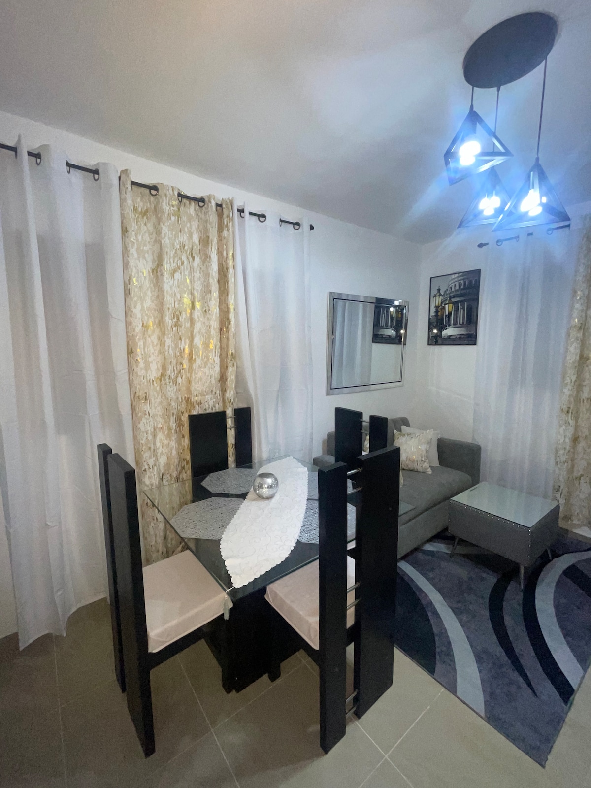A dining area features a glass table surrounded by four black chairs, with a simple centerpiece placed on the table. A light fixture with several bulbs illuminates the space, while patterned curtains frame a large window. A cozy sofa adds comfort to the area.