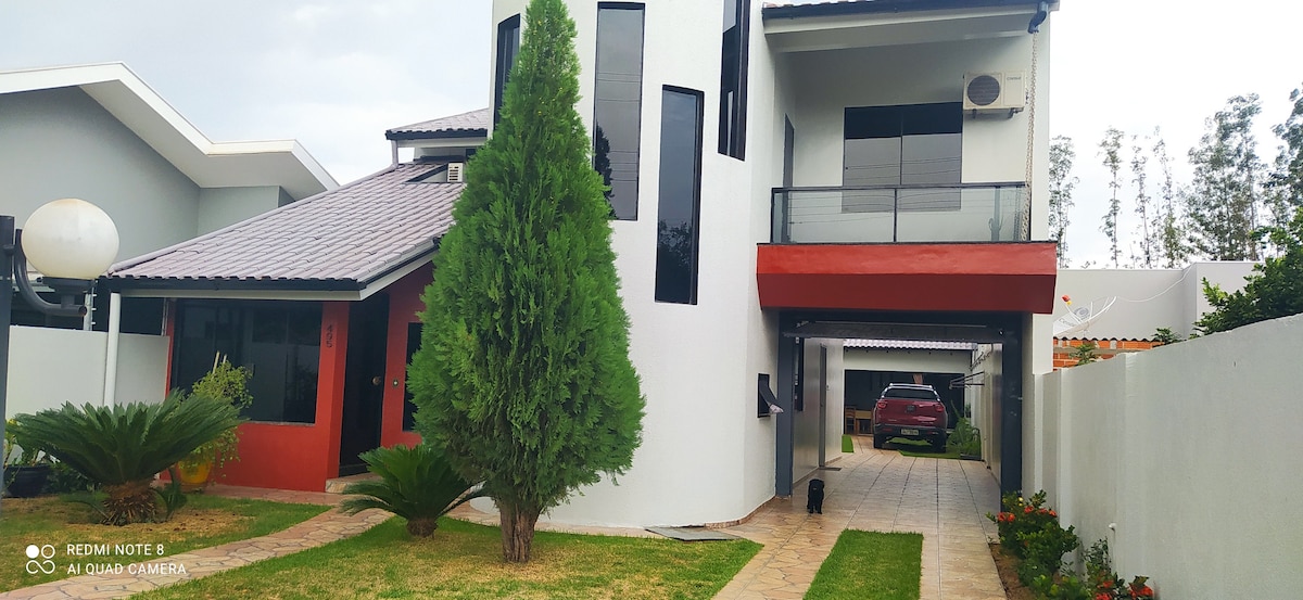 A modern two-story house features a combination of red and white walls, with expansive glass windows allowing natural light. A well-maintained lawn with small shrubs and palm plants enhances the curb appeal. A spacious driveway is visible, leading to a covered parking area.