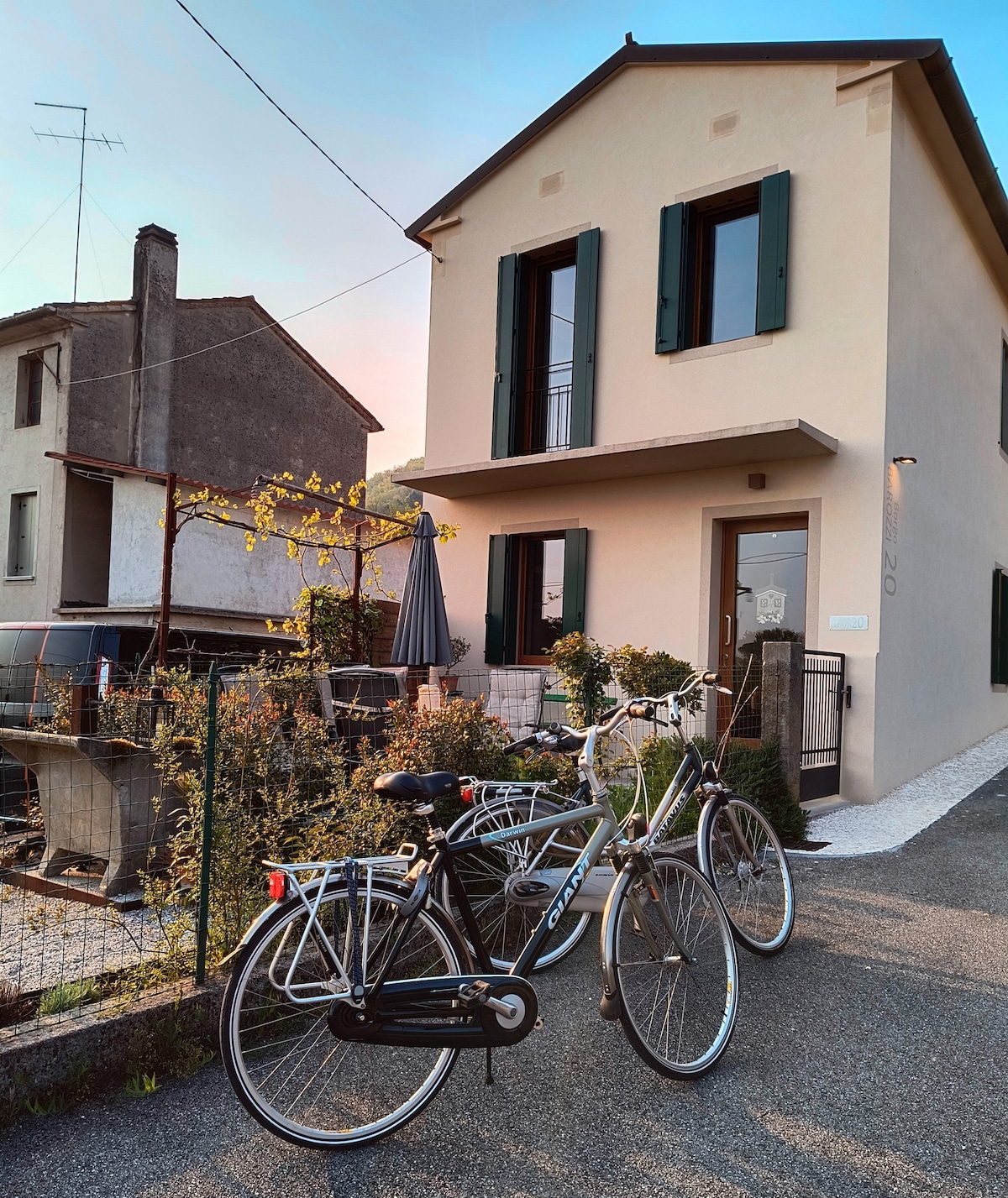 A quaint two-story house, featuring green shutters, stands at the edge of a quiet path. Two bicycles are parked in front, with a small garden visible, including plants and a table with an umbrella. Soft lighting highlights the warm exterior under the evening sky.