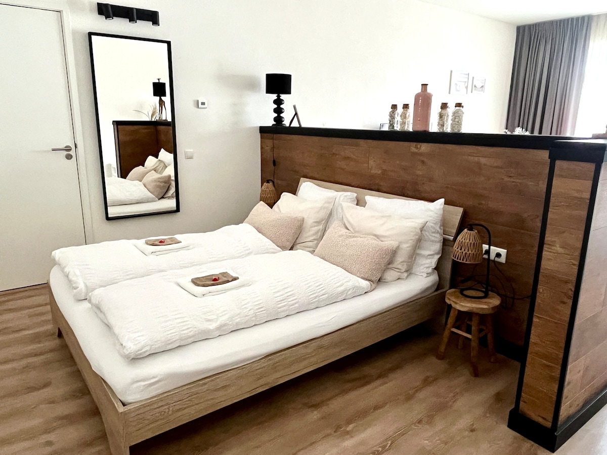 The sleeping area features a spacious bed with soft linens and decorative pillows. A wooden partition adds privacy, while a full-length mirror reflects the soft, neutral tones of the room. A small wooden chair and decorative elements enhance the warm ambiance.