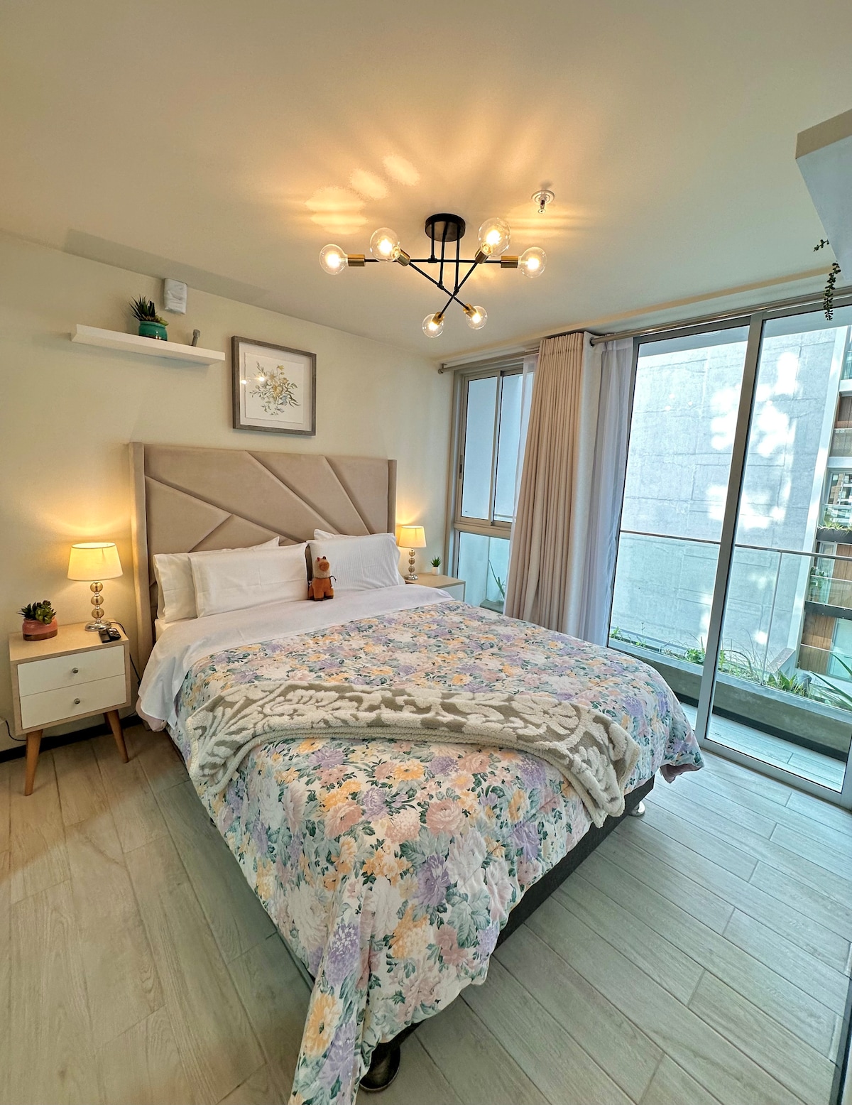 A bright bedroom features a spacious bed adorned with a floral blanket and decorative cushions. Two bedside tables with lamps are positioned beside the bed. Large windows allow natural light to fill the room, showcasing a balcony view with greenery.