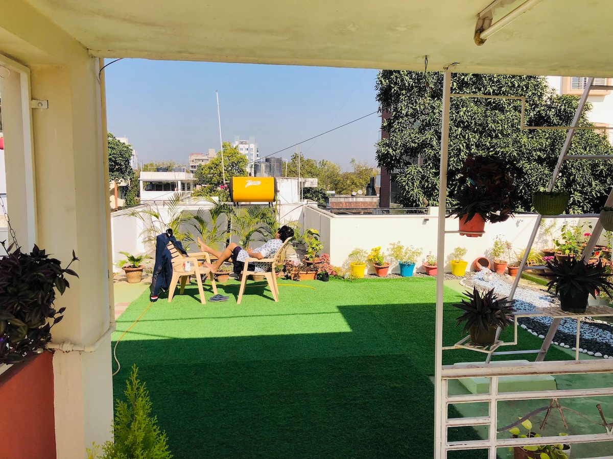 A spacious outdoor terrace is shown, featuring green artificial turf and various potted plants. Relaxing seating arrangements, including chairs, are visible under a clear blue sky. The area is framed by a railing and has easy access through the building structure.