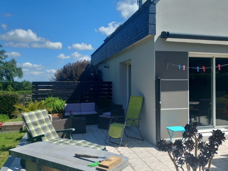 An outdoor terrace features modern lounge furniture and sun loungers arranged on tiled flooring. A green and white patterned chaise lounge rests alongside a small blue table. Nearby, a low seating area with cushioned chairs complements the natural setting, enhancing the peaceful atmosphere.