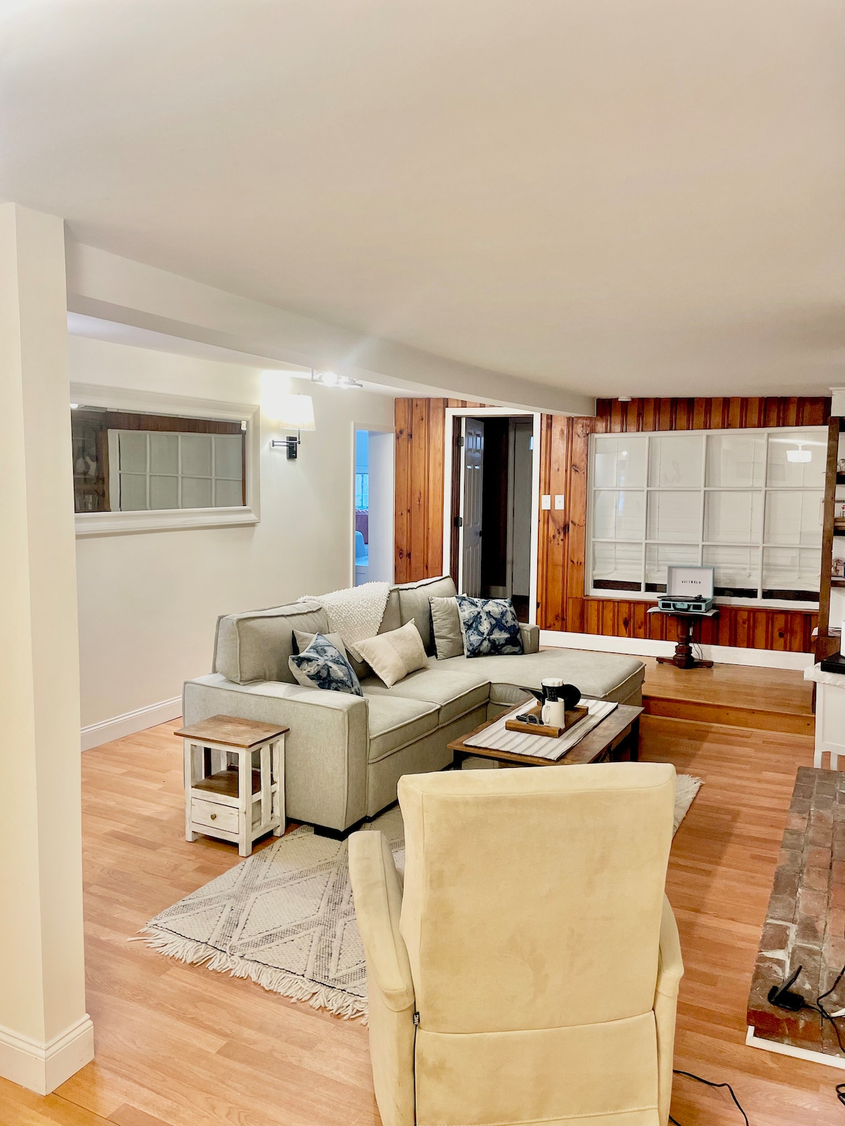 A spacious living area is arranged with a comfortable couch featuring decorative pillows. A coffee table sits in front, accompanied by a small side table. Natural light fills the room, highlighting the wooden accents on the walls and the soft texture of the area rug.