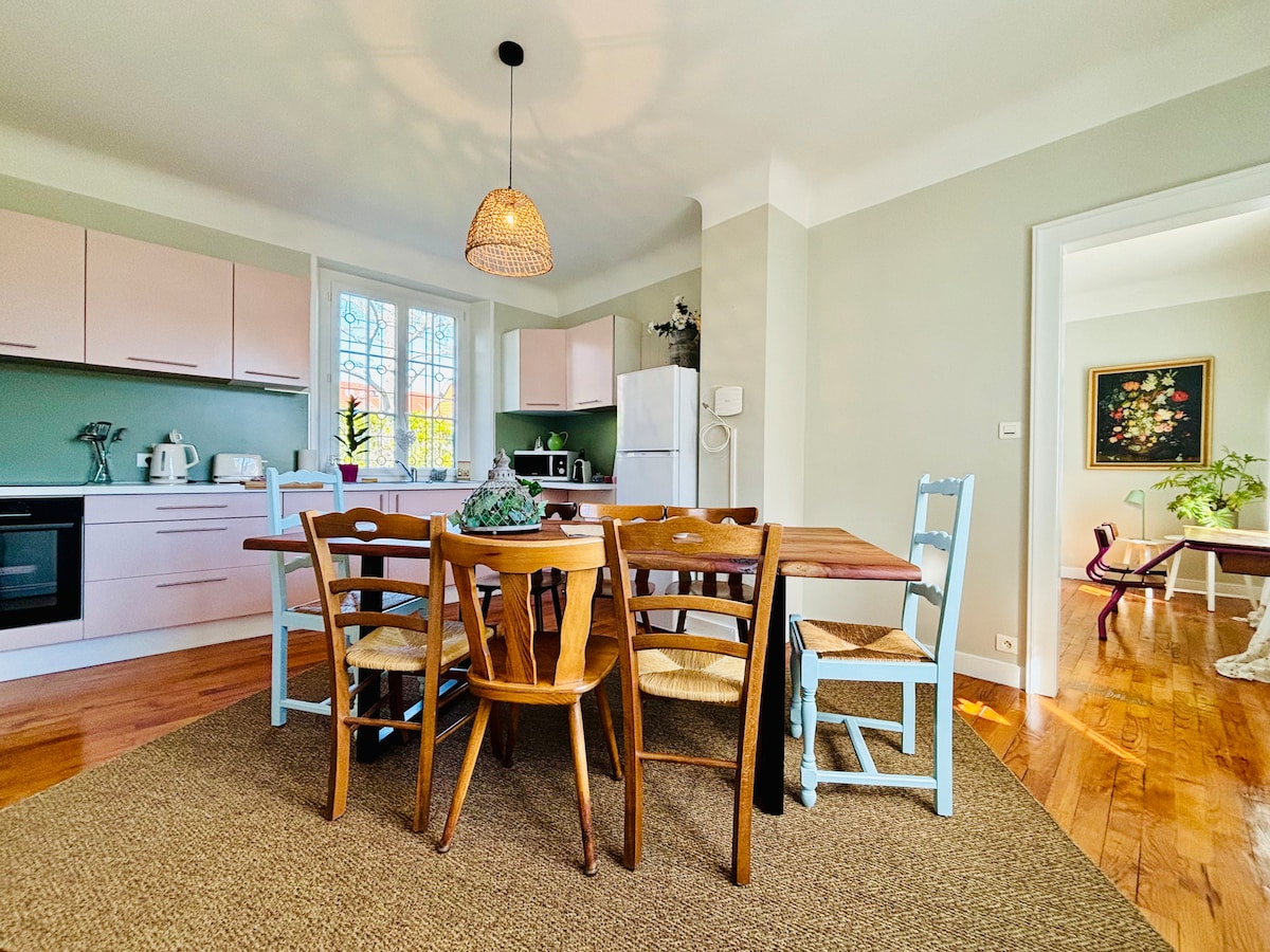 The kitchen and dining area features a wooden dining table surrounded by a mix of chairs, including light blue and natural wood. A pendant light hangs from the ceiling, and large windows allow natural light to illuminate the space. The kitchen is well-equipped with modern appliances.