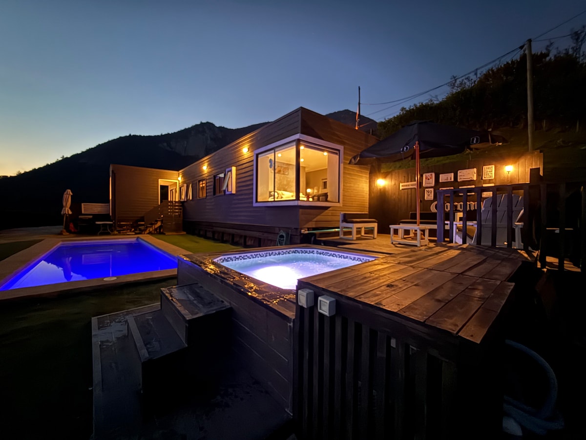 An outdoor area is illuminated at dusk, featuring a pool and a jacuzzi. Wooden decking surrounds both water features, creating a space for relaxation. The structure showcases large windows, allowing for views of the surrounding mountains, while warm lighting highlights the inviting atmosphere.