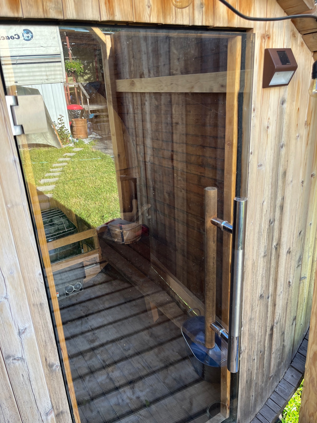 A wooden sauna with a glass door invites relaxation, featuring a minimalist interior lined with wood. A small bucket is positioned on the floor, and the space is framed by natural light filtering through the glass.