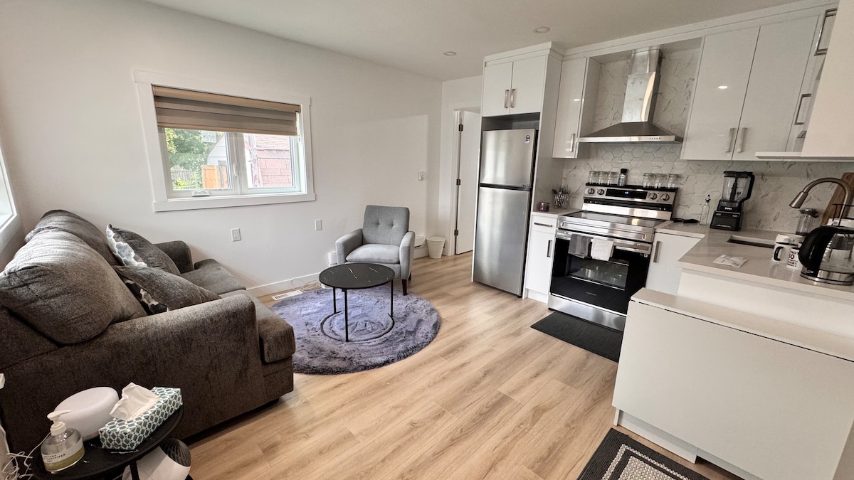 A contemporary living area features a comfortable gray sofa and a small round table. The modern kitchen showcases stainless steel appliances and sleek cabinets. An accent chair is positioned near a large window, providing natural light to the space. Wooden flooring complements the neutral color scheme.