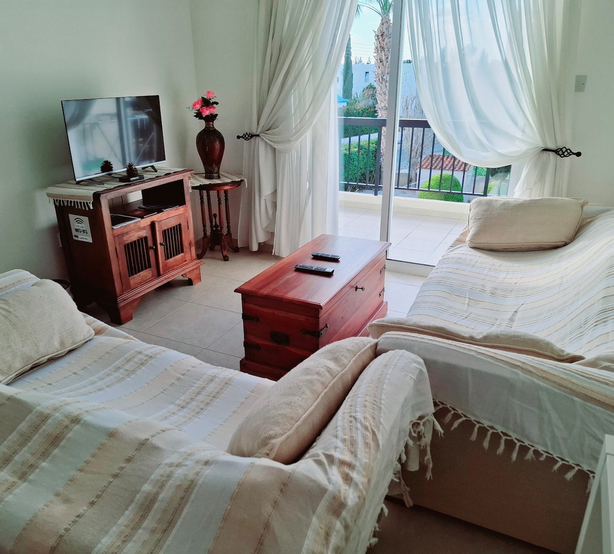 A living area is shown, featuring two light-colored couches adorned with soft throws. A wooden coffee table sits in the center, and a wooden TV stand holds a flat-screen television. Sheer white curtains allow natural light to illuminate the space, with a glimpse of the balcony visible through the open doors.