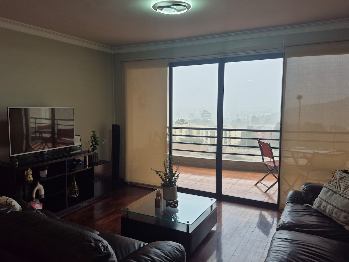 A spacious living area features a large television on a low black entertainment unit. Floor-to-ceiling sliding glass doors offer a view of the city, while a small balcony with a chair provides an outdoor seating option. Natural light filters through sheer curtains.