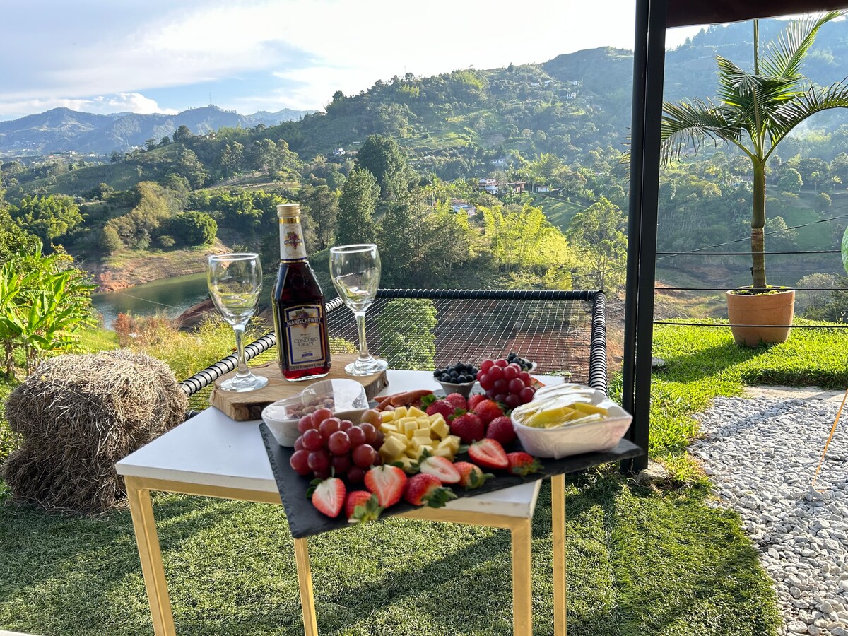 A small table is set outdoors, featuring a variety of fresh fruits including strawberries, grapes, and slices of tropical fruits. Two glasses and a bottle of wine sit atop the table, with a scenic view of the lush green hills and lake in the background.